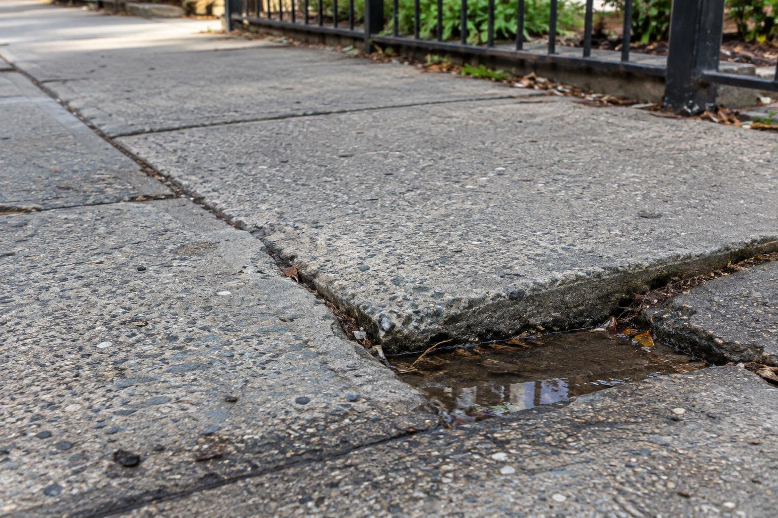 Displaced sidewalk crack and low concrete edge holding a small puddle.